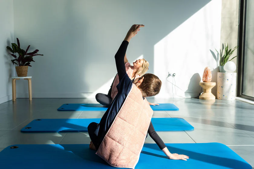 Two people sit on blue yoga mats indoors, stretching one arm upward while wearing light pink shawls. Sunlight streams through a large window, and potted plants decorate the minimalist space.
